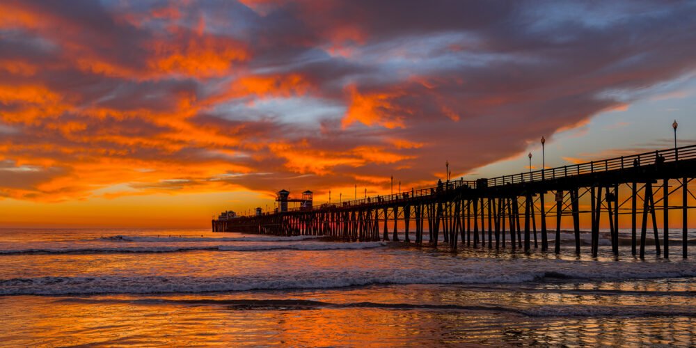 A colorful winter sunset illuminates the sky over Oceanside Pier in this panoramic image along the California Coast in northern San Diego