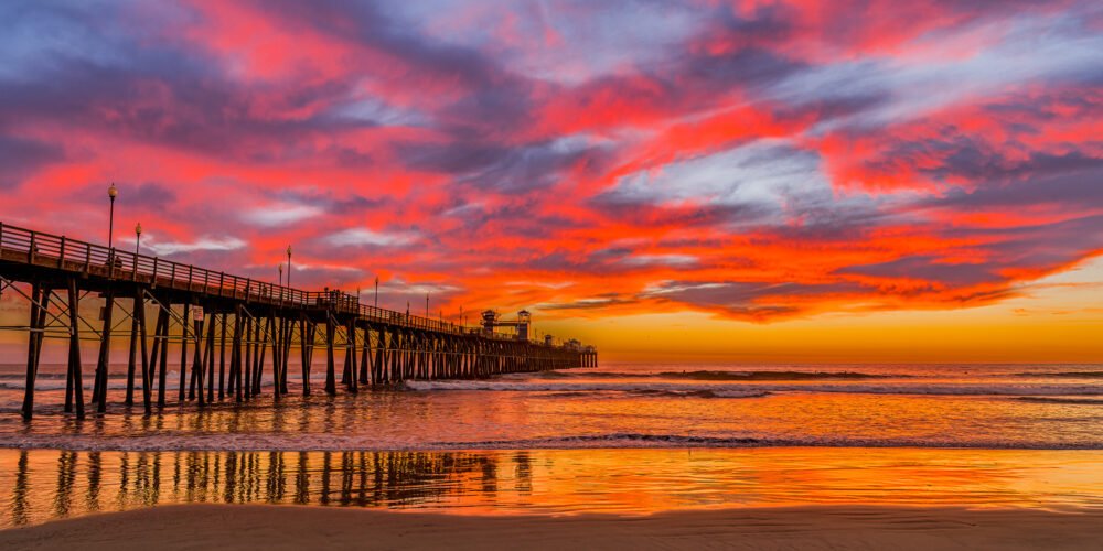 A colorful winter sunset punctuates the sky as Oceanside Pier presides over the calm surf in this panoramic image along the California Coast
