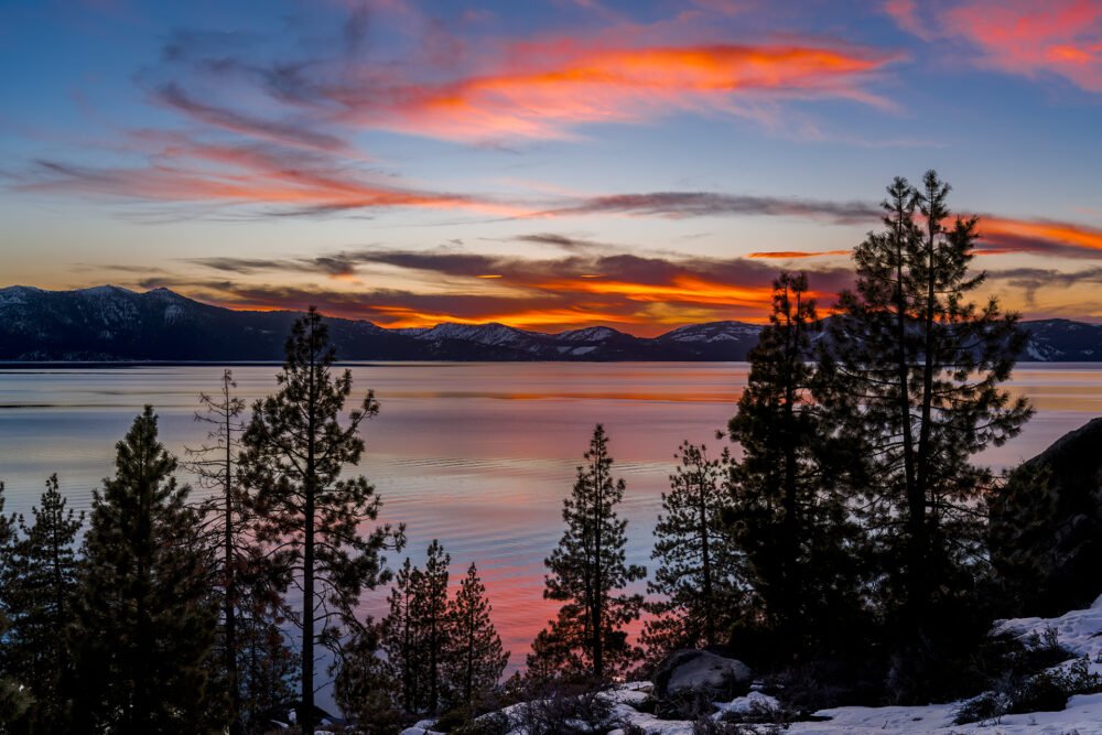 Pine trees and mountains frame this scene at Lake Tahoe with sunset reflecting in the lake.
